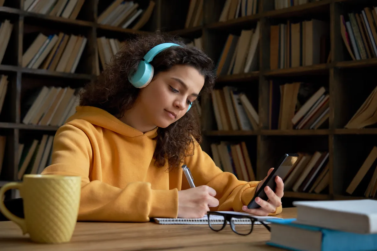 mujer en una biblioteca escuchando música y anotando en una libreta